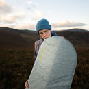 A person wearing glasses and a blue knit hat inflates the Therm-a-Rest Trail Lite Sleeping Pad outdoors, with hills and a cloudy sky in the background—an ideal setup for lightweight backpacking adventures.