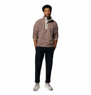 A man faces forward against a white background, wearing the Columbia Mens Helvetia Half Snap Fleece in light brown, paired with dark pocketed pants and light sneakers.