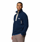 A man wears the Columbia Mens Helvetia Half Snap Fleece in navy blue with a beige snap-button collar and zippered chest pocket, standing against a plain white background with one hand in his pocket, looking off to the side.