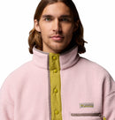 A man with long brown hair wears the Columbia Mens Helvetia Half Snap Fleece—a retro light pink Sherpa pullover with a high collar, yellow snaps, and logo patch—from the Icons collection, standing against a white background.
