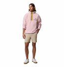 A man wears the Columbia Mens Helvetia Half Snap Fleece in light pink with a partial yellow zipper, paired with beige shorts and white sneakers, standing against a plain white background.