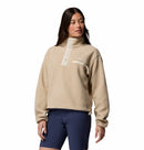 A woman in a beige Columbia Womens Helvetia II Cropped Sherpa Half Snap Fleece with a high collar, snap buttons, and chest pocket, paired with navy blue shorts, stands slightly turned against a plain white background.