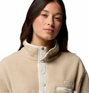 A young woman with long brown hair wears the Columbia Womens Helvetia II Cropped Sherpa Half Snap Fleece, a PFAS-free, water-repellent beige jacket with a high collar, standing against a white background and looking confidently at the camera.