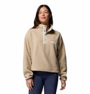 A woman wears the Columbia Womens Helvetia II Cropped Sherpa Half Snap Fleece, featuring a button-up collar and water-repellent chest pocket, paired with dark blue pants against a plain white background.