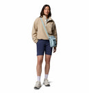 A person stands against a white background wearing the Columbia Womens Helvetia II Cropped Sherpa Half Snap Fleece in beige, navy shorts, white socks, sneakers, and a light blue crossbody bag.