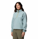 A woman wears the Columbia Womens Helvetia II Cropped Sherpa Half Snap Fleece in light blue with a zippered chest pocket and olive green pants, posed against a plain white background.