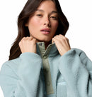 A woman with long brown hair wears the Columbia Womens Helvetia II Cropped Sherpa Half Snap Fleece in light blue, holding the collar with both hands and looking forward with a neutral expression against a plain background.