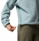 A person wearing olive green pants and a light blue Columbia Womens Helvetia II Cropped Sherpa Half Snap Fleece is adjusting the drawstring at the pullover’s hem.