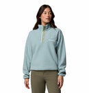 A woman with long dark hair, dressed in olive green pants and the Columbia Womens Helvetia II Cropped Sherpa Half Snap Fleece—light blue, high collar, snap buttons, and chest pocket—stands against a plain white background.