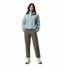 A woman with long brown hair faces forward, wearing the Columbia Womens Helvetia II Cropped Sherpa Half Snap Fleece, olive green pants, and white sneakers. She poses with one hand in her pocket against a white background.