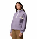 A woman wears the Columbia Womens Helvetia II Cropped Sherpa Half Snap Fleece in light purple, featuring a high collar, buttoned placket, and zippered chest pocket, paired with tan pants against a white background.