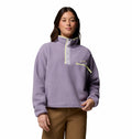 A woman with long dark hair poses against a white background wearing Columbias Womens Helvetia II Cropped Sherpa Half Snap Fleece in light purple, featuring a high collar, button-up placket, and zippered chest pocket, paired with brown pants.