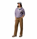 A person wearing a light gray cap, Columbia Womens Helvetia II Cropped Sherpa Half Snap Fleece in lavender, light blue shirt, brown pants, and hiking shoes stands with one hand in their pocket against a white background.