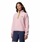 A woman wears the Columbia Womens Helvetia II Cropped Sherpa Half Snap Fleece in light pink with a zipper and button placket, paired with navy shorts, against a white background. The fleece is PFAS-free and water-repellent.