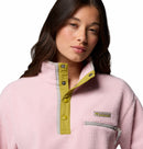 A woman with long brown hair wears the Columbia Womens Helvetia II Cropped Sherpa Half Snap Fleece in light pink, featuring a yellow and gray snap-button collar and zippered chest pocket, posing against a white background.