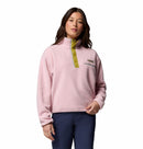 A woman with long dark hair and a neutral expression wears the Columbia Womens Helvetia II Cropped Sherpa Half Snap Fleece in light pink with green buttons and collar, paired with dark blue pants against a white background.