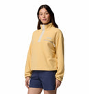 A woman wears the Columbia Womens Helvetia II Cropped Sherpa Half Snap Fleece in light yellow, featuring a PFAS-free water-repellent finish and snap buttons, paired with navy shorts, standing against a plain white background.