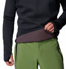 A person wearing the Columbia Mens Crystal Leaf Omni-Heat Helix Technical Half Zip Fleece in black and green pants lifts the shirt, revealing a stretchy waistband with a pink and black polka dot pattern.