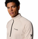A man with short curly hair wears a light beige Columbia Mens Crystal Leaf Omni-Heat Helix Technical Half Zip Fleece, featuring a black zipper, chest logo, and a small black sleeve graphic, standing against a white background.