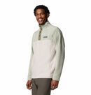 A smiling man wears the Columbia Mens Steens Mountain™ II Half Snap Fleece, a light green and cream PFAS-free pullover with a standing collar and brown pants, standing against a plain white background—ideal for everyday wear.