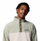 A man with short curly hair wears the Columbia Mens Steens Mountain™ II Half Snap Fleece, a PFAS-free pullover in light green and off-white, featuring a high collar and snap buttons. He looks slightly to the side with a soft smile.
