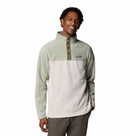 A man smiles wearing the Columbia Mens Steens Mountain™ II Half Snap Fleece in light green and cream, featuring a high collar and snap placket, paired with olive pants against a white background.