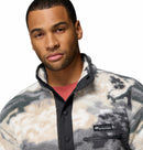 A man with short hair wears Columbias Mens Helvetia II Printed Half Snap Fleece in a black, white, and beige pattern over a red shirt. The pullover features a black snap collar and a logo patch on the chest.