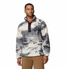A man with short hair wears the Columbia Mens Helvetia II Printed Half Snap Fleece, featuring a high collar, snap buttons, and chest pocket. He pairs it with brown pants and stands against a plain white background.