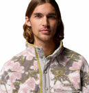 A person with long brown hair wears the Columbia Mens Helvetia II Printed Half Snap Fleece, featuring a soft textured pink, green, and white pattern. The pullover has a high collar and button details. They face forward against a white background.