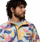 A man with curly hair and a beard wears the Columbia Mens Helvetia II Printed Half Snap Fleece, featuring a pastel abstract pattern, yellow accents, and the Columbia logo on the chest, looking sideways against a white background.