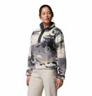 A woman stands against a plain white background, wearing Columbias Womens Helvetia II Cropped Sherpa Half Snap Printed Fleece in gray, black, and white print with a buttoned collar, paired with light-colored pants, looking slightly to the side.