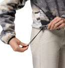 A woman in light-colored pants tightens the elastic drawcord at the hem of a Columbia Womens Helvetia II Cropped Sherpa Half Snap Printed Fleece jacket against a white background.