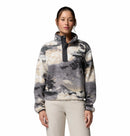 A woman wears the Columbia Womens Helvetia II Cropped Sherpa Half Snap Printed Fleece in grey and white, featuring a high collar and snap buttons, paired with light-colored pants. She stands against a plain white background with her brown hair tied back.