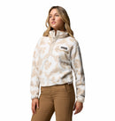 A woman wears the Columbia Womens Helvetia II Cropped Sherpa Half Snap Printed Fleece in light beige with a floral pattern and tan pants, standing against a white background and gazing to the side with a neutral expression.