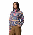 A person wears the Columbia Womens Helvetia II Cropped Sherpa Half Snap Printed Fleece, featuring a colorful design with a pink snap collar, paired with brown pants and one hand in their pocket against a white background.