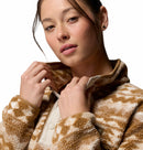 A young woman with fair skin and brown hair pulled back wears the Columbia Womens Helvetia II Cropped Sherpa Half Snap Printed Fleece, holding the collar with both hands. She has light makeup, small triangular earrings, and stands against a white background.