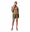 A woman stands against a white backdrop wearing the Columbia Womens Helvetia II Cropped Sherpa Half Snap Printed Fleece in tan and white, with olive shorts and blue sandals. She touches her neck and appears relaxed.