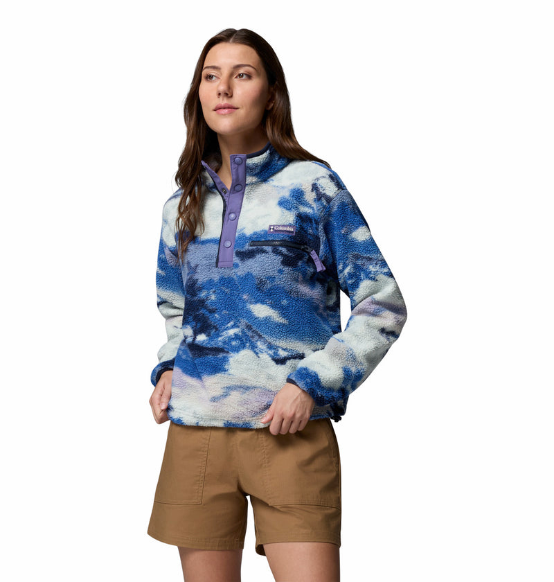A woman models the Columbia Womens Helvetia II Cropped Sherpa Half Snap Printed Fleece in blue and white, featuring a purple buttoned collar, paired with tan shorts. She stands hands-in-pockets against a plain white background.