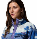 A woman with long brown hair wears the Columbia Womens Helvetia II Cropped Sherpa Half Snap Printed Fleece, featuring a blue and white PFAS-free design, purple snap-up collar, and logo on the chest, against a white background.