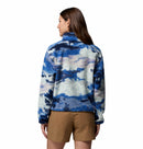 A woman with long brown hair wears tan shorts and the Columbia Womens Helvetia II Cropped Sherpa Half Snap Printed Fleece in a blue, white, and purple cloud print. She stands facing away from the camera against a plain white background.