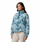 A woman models the Columbia Womens Helvetia II Cropped Sherpa Half Snap Printed Fleece, featuring a blue and white print, high collar, and snap buttons. She pairs it with light-colored pants against a white background.