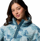 A woman with long brown hair wears the Columbia Womens Helvetia II Cropped Sherpa Half Snap Printed Fleece in light blue, featuring a high collar, snap buttons, and a zippered chest pocket. She looks slightly to the side and smiles softly.