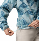 A person adjusts the hem drawstring of a Columbia Womens Helvetia II Cropped Sherpa Half Snap Printed Fleece in blue and white, paired with light beige pants. Only their torso and hands appear against a white background.