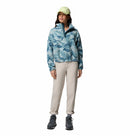 A woman wears the Columbia Womens Helvetia II Cropped Sherpa Half Snap Printed Fleece, a light green cap, cream pants, and hiking boots, standing facing forward against a white background.