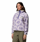 A woman wears the Columbia Womens Helvetia II Cropped Sherpa Half Snap Printed Fleece in light purple and white, featuring a high collar and snap buttons, paired with olive green pants against a plain white background.