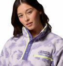 A woman with wavy brown hair wears the Columbia Womens Helvetia II Cropped Sherpa Half Snap Printed Fleece, featuring a light purple and white design, high collar, purple snaps, chest zip pocket, and the Columbia logo.