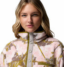 A young woman with long blonde hair models the Columbia Womens Helvetia II Cropped Sherpa Half Snap Printed Fleece, a PFAS-free jacket with pink, white, and green abstract prints, zippered chest pocket, and logo, against a white background.