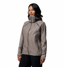 A woman with medium-length dark hair wears a Columbia Womens Reign No Shine Waterproof Jacket in light gray and dark pants, standing against a plain white background, prepared for optimal rain protection.