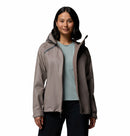A woman with medium-length dark hair is wearing the Columbia Womens Reign No Shine Waterproof Jacket in light gray over a light blue shirt and dark pants, standing against a plain white background.