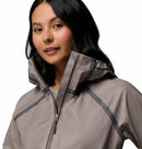 A woman with dark hair wears the Columbia Womens Reign No Shine Waterproof Jacket, featuring a high collar and hood, standing against a plain white background.